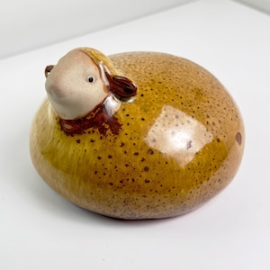 May include: A ceramic sheep figurine with a tan face and ears, and a speckled, rounded body in shades of brown and yellow. The sheep has a small, rounded body and is resting on a white surface. The figurine is a decorative item.