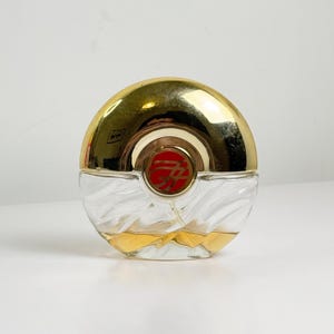 Peut inclure: Un flacon de parfum rond en verre avec une base texturée en spirale et un bouchon en métal doré brillant. Le centre du bouchon présente un emblème rouge circulaire avec un logo doré stylisé. Le flacon contient un liquide ambré clair.
