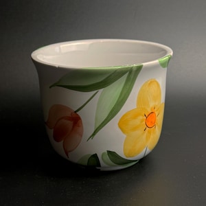 May include: White ceramic plant pot with a hand-painted floral design. It features yellow and orange flowers with green leaves. The rim is painted green. A decorative item for plants or home decor.