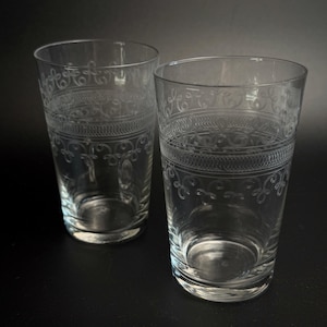 Op de afbeelding: Twee heldere glazen drinkbekers met een geëtst ontwerp rond de bovenrand. Het patroon bestaat uit een horizontale band van fijne verticale lijnen, omrand door twee rijen delicate, herhalende krulmotieven gegraveerd in het glas.