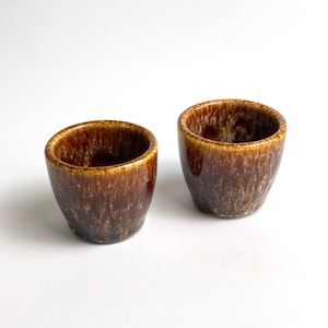 Pode incluir: Duas pequenas chávenas de cerâmica castanha com acabamento brilhante. As chávenas têm formato arredondado e textura salpicada, com uma borda castanha mais clara. As chávenas estão sobre um fundo branco.