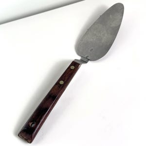 May include: A vintage pie server. It has a stainless steel blade and a dark brown wooden handle. The handle is secured with two brass rivets and has a hole at the end. The server is designed for cutting and serving pies or cakes.