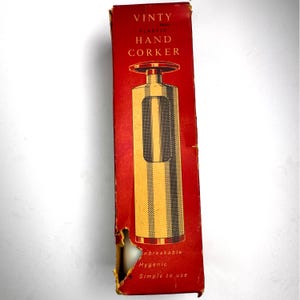 May include: A vintage red and gold hand corker in its original packaging. The box reads "VINTY PLASTIC HAND CORKER." The corker is gold with a black and gold striped design. The box has a tear on the side.
