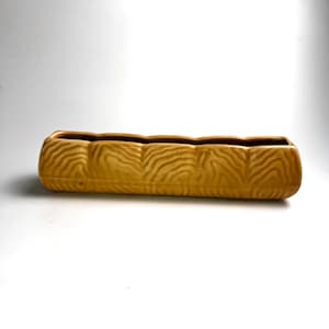 Puede incluir: Una jardinera rectangular de cerámica amarillo mostaza con un patrón texturizado en forma de onda. La jardinera tiene una forma larga, similar a un abrevadero, con una serie de hendiduras curvas a lo largo del borde superior. El interior es de color marrón oscuro.