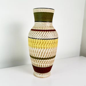 May include: A decorative vase with a woven texture and a pattern of horizontal bands. The vase features colours such as olive green, yellow, and burgundy against a cream backdrop. It has a narrow neck and a wide base, typical of a floor vase.