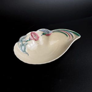 May include: A cream-colored, ceramic serving dish with a floral design. The dish has a scalloped edge and is decorated with pink, blue, and green painted flowers and leaves. The dish is on a black surface.