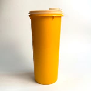 Può includere: Un bicchiere alto di plastica gialla con coperchio abbinato e chiusura a ribalta beige. Il bicchiere è cilindrico e sembra vuoto. Il coperchio si adatta perfettamente sopra e il ribaltabile è in posizione chiusa. Lo sfondo è bianco.