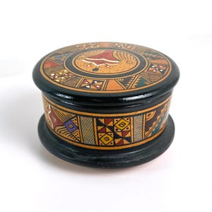 May include: A round, black wooden box with a lid. The lid and sides are decorated with colourful geometric patterns and shapes in red, yellow, blue, and brown. The box sits on a black base.