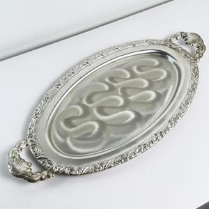 May include: A silver oval serving tray with decorative detailing. The tray has a patterned border and shell-shaped handles. The interior has a wavy design. A classic piece for serving food or displaying items, measuring approximately 40cm in length.