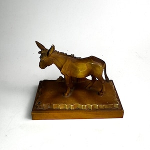 Puede incluir: Figurilla de burro de madera tallada con un cuerno, de pie sobre una base rectangular. El burro está tallado con textura visible y es de color marrón dorado cálido. La base tiene un borde decorativo.