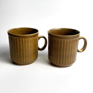 May include: Two matching ceramic mugs in a deep olive-brown color. Each mug features a cylindrical body with vertical ribbed detailing along the exterior surface, a smooth rim, and a curved handle attached to the side for holding the vessel.