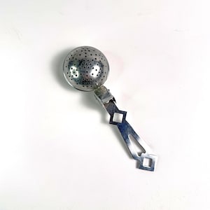 May include: A silver-toned metal tea infuser featuring a spherical, perforated mesh head attached to a flat, elongated handle. The handle is designed with a series of geometric cutouts, including two square shapes and a central diamond-like opening.