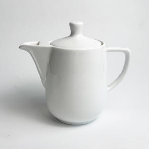 Puede incluir: Una tetera de cerámica blanca con tapa y asa. La tetera tiene un pico para verter y un cuerpo redondeado. La tapa tiene un pequeño pomo en la parte superior. La tetera está diseñada para preparar y servir té.