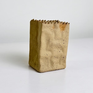 May include: A beige ceramic vase shaped like a crumpled paper bag with a speckled texture and a wavy, serrated top edge. The surface features irregular folds and indentations, with a small patch of orange-brown discolouration near the top rim.