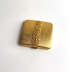 May include: A square gold-toned metal compact case with a textured, brushed finish. A vertical strip of intricate, openwork filigree detailing runs down the center of the lid. The compact features a small metal latch on the front edge.