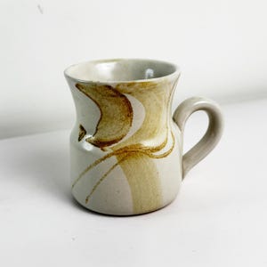 May include: A cream-coloured ceramic mug with a curved handle. The mug has brown abstract designs and a slightly flared rim. Suitable for hot or cold drinks. Capacity is approximately 300 ml.