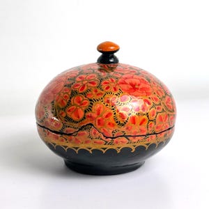 Puede incluir: Una caja decorativa redonda con tapa y diseño floral. La caja es principalmente roja con patrones florales naranjas y dorados sobre un fondo negro. La base es negra con un borde dorado, y la tapa tiene un pequeño pomo naranja.