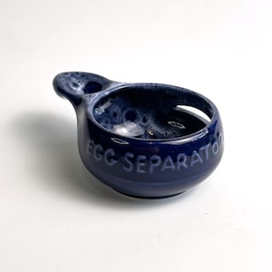 Könnte beinhalten: Ein marineblauer Eiertrenner aus Keramik mit Griff und Ausguss. Die Worte "EGG SEPARATOR" sind auf der Seite geprägt. Der Innenraum hat eine gesprenkelte, hellere blaue Glasur.