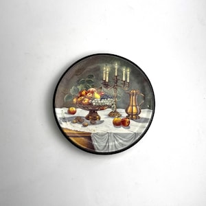 Puede incluir: Un plato decorativo redondo con una naturaleza muerta. La imagen presenta un cuenco de fruta, un candelabro con velas encendidas y una jarra dorada. El plato tiene un borde negro y está sobre un fondo oscuro.