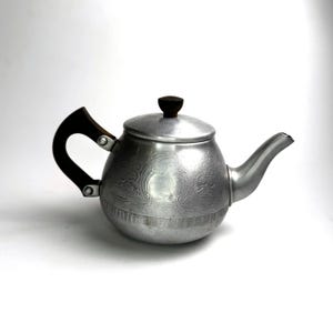 May include: A vintage silver teapot with a dark wooden handle and lid knob. The teapot features an embossed floral design on the body and a curved spout. The handle is attached with metal rivets.