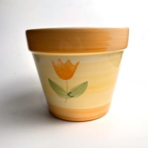 Könnte beinhalten: Keramik-Blumentopf mit handgemaltem Tulpen-Design. Der Topf hat ein gelb-oranges Farbschema mit einer Tulpe in Orange und grünen Blättern. Der Rand und der Boden sind dunkler orange. Der Topf ist zum Bepflanzen von Blumen bestimmt.