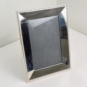 May include: A silver-colored picture frame with a rectangular opening. The frame has a beaded border and a reflective surface. The frame is standing upright on a white surface.