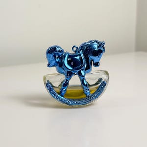 Könnte beinhalten: Ein kleines Parfümfläschchen aus Glas in Form eines Schaukelpferdes mit metallisch blauer Oberfläche. Der klare Glassockel enthält eine gelbe Flüssigkeit, und die Pferdefigur verfügt über eine kleine Öse auf dem Rücken zum Aufhängen.
