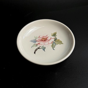 May include: A small, off-white ceramic dish with a hand-painted floral design. The flower is pink and white with green leaves and a brown stem. The dish is round with a slightly raised edge.