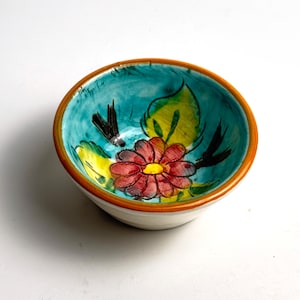 May include: A small, hand-painted ceramic bowl featuring a floral design. The interior is turquoise with a red flower, yellow leaves, and black accents. The rim is brown, and the exterior is white.