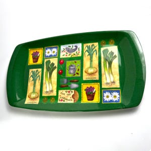 Könnte beinhalten: Ein rechteckiges, grünes Serviertablett mit Gartenthema. Das Tablett zeigt Illustrationen von Zwiebeln, Lauch, Blumen, Gartengeräten und den Text "FOR THE GARDENER". Die Illustrationen sind vor einem cremefarbenen Hintergrund.