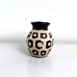 Puede incluir: Un pequeño jarrón decorativo con cuerpo color crema y borde marrón oscuro. El jarrón presenta un patrón repetitivo de cuadrados marrones con círculos blancos en el interior. El jarrón mide unos 10 cm de altura.