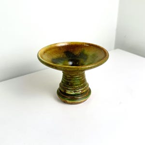 Puede incluir: Un cuenco de cerámica con pedestal, con una parte superior ancha y poco profunda y una base texturizada y acanalada. El cuenco presenta una mezcla de tonos terrosos, incluyendo tonos de marrón, verde y amarillo. La pieza de cerámica hecha a mano se utiliza probablemente con fines decorativos.