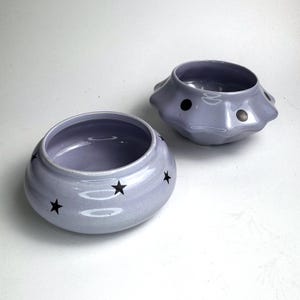 Könnte beinhalten: Zwei hellviolette Keramikschalen. Eine Schale ist rund mit schwarzen Sterndesigns. Die andere Schale hat eine einzigartige Form mit schwarzen Punktdesigns. Beide Schalen haben eine glänzende Oberfläche und sind leer.
