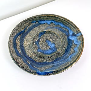 Può includere: Un piatto in ceramica fatto a mano con un design a spirale unico. Il piatto presenta una miscela di colori blu, grigio e beige, con una finitura lucida. La forma circolare e la superficie strutturata aggiungono al suo fascino artistico.