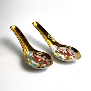 Könnte beinhalten: Zwei chinesische Suppenlöffel aus Keramik mit goldfarbenen Griffen und Rändern. Die Löffel haben weiße Schalen mit einem roten Drachendesign und grünen und gelben Akzenten. Auf weißem Hintergrund.