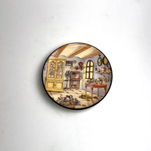 Puede incluir: Plato decorativo con una ilustración detallada de un interior de cocina acogedor. La escena incluye una chimenea, una mesa, un armario y varios artículos de cocina. El plato tiene un borde negro y un acabado brillante.