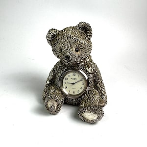 May include: A silver-toned teddy bear ornament with a clock face. The clock has a white face with black numerals and hands. The bear is in a seated position, with its legs extended and arms at its sides.
