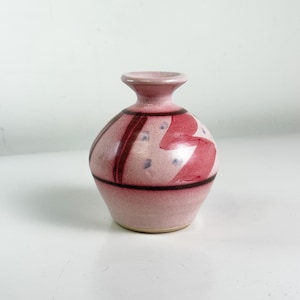 May include: A small, pink ceramic vase with a rounded body and a narrow neck. The vase features dark red stripes and abstract red and purple painted designs. The vase is approximately 4 inches tall.