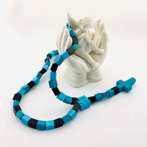May include: A necklace made of blue, black, and turquoise square blocks, with a cross-shaped pendant. The necklace is displayed in front of a white ceramic angel figurine.