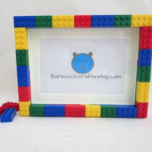 Puede incluir: Un marco de fotos blanco decorado con bloques de construcción de juguete de colores rojo, azul, verde y amarillo. El marco tiene un interior blanco con una ilustración de oso de dibujos animados y el texto "Baresockcrafts.etsy.com".
