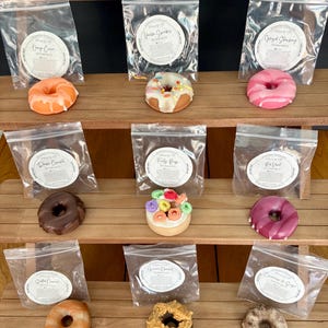 May include: Nine different donut-shaped candles arranged on a wooden shelf. Each candle is in a clear plastic bag with a label that includes the scent name and a small graphic of a donut. The scents include: Orange Cream, Vanilla Sprinkles, Glazed Strawberry, Dark Coconut, Fruity Rings, Red Velvet, Salted Caramel, Peanut Butter, and Cinnamon & Sugar.