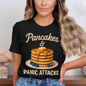 May include: Black t-shirt with the words "Pancakes & Panic Attacks" in a whimsical font. The shirt features a stack of pancakes with butter and syrup, and blueberries on a plate.