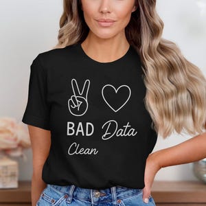 Op de afbeelding: Zwart T-shirt met de tekst "BAD Data Clean" en een vredesteken en hart in wit. Het shirt heeft een ronde hals en is gemaakt van een zacht materiaal.