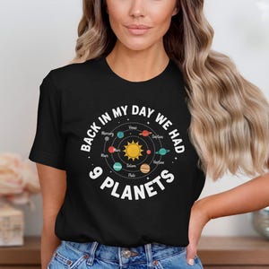 May include: Black t-shirt featuring a solar system graphic and the text "BACK IN MY DAY WE HAD 9 PLANETS". The design includes the sun and planets in a circular layout. The shirt is worn by a person.