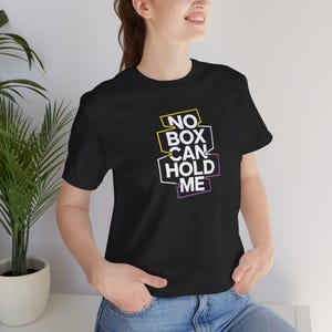 Puede incluir: Camiseta negra con el texto "NO BOX CAN HOLD ME" en letras blancas con contornos amarillos y morados.