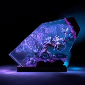 Puede incluir: Escultura decorativa de resina con una criatura blanca posada en la rama de un árbol. La escultura está encerrada en un bloque transparente en ángulo con patrones arremolinados morados y azules. La base es un rectángulo de madera oscura.