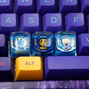 Puede incluir: Primer plano de un teclado mecánico con teclas moradas. Tres teclas personalizadas presentan logotipos de equipos para Chelsea, Inter de Milán y Manchester City. La tecla "ALT" es amarilla, las otras son moradas.