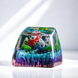 Pode incluir: Uma tecla de resina colorida, com miniaturas de um personagem vermelho e um branco, num cenário de paisagem azul, verde e roxa. A tecla tem formato quadrado e acabamento transparente e brilhante.