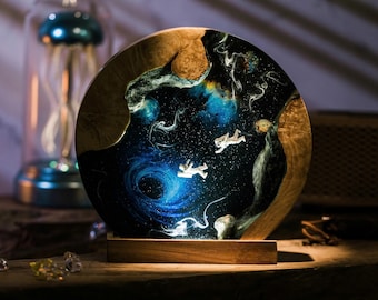 Puede incluir: Una base de lámpara redonda de madera con una incrustación de resina que representa una galaxia giratoria con dos astronautas flotando en el espacio. La base de la lámpara está iluminada desde el interior, proyectando un brillo cálido.