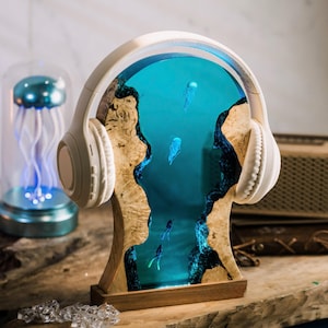 May include: White headphones with a wooden stand featuring a blue resin insert with jellyfish and fish. The stand is made of wood with a natural wood grain.